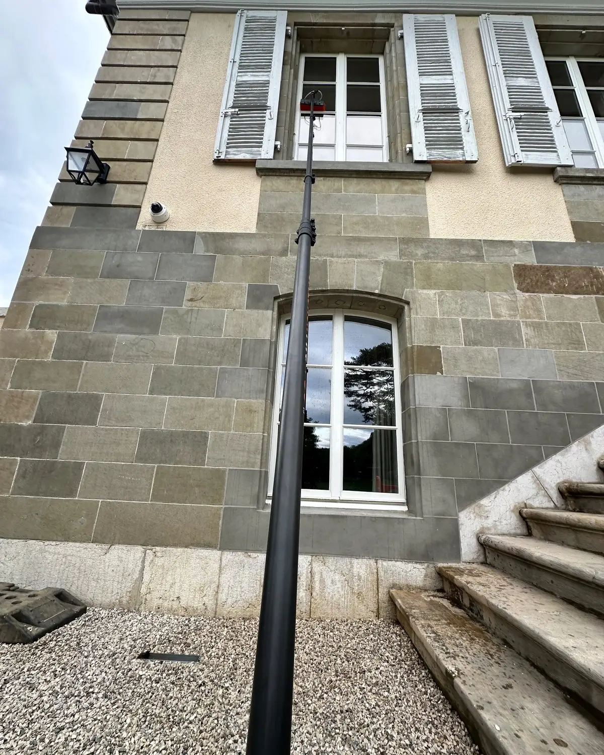 Perche télescopique atteignant une fenêtre en étage d'une maison en pierre, près d'un perron